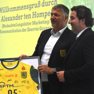 Alexander ten Hompel von MARK-E und Geschäftsführer Fynn Holpert präsentieren beim Saisonauftakt des VfL Eintracht Hagen ein gerahmtes Mannschaftstrikot.